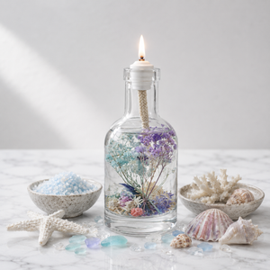 Lit Pisces oil candle: a clear glass bottle of pastel blue and lilac dried flowers with a burning wick, on white marble beside bowls of blue sea salt, coral, a starfish and shells
