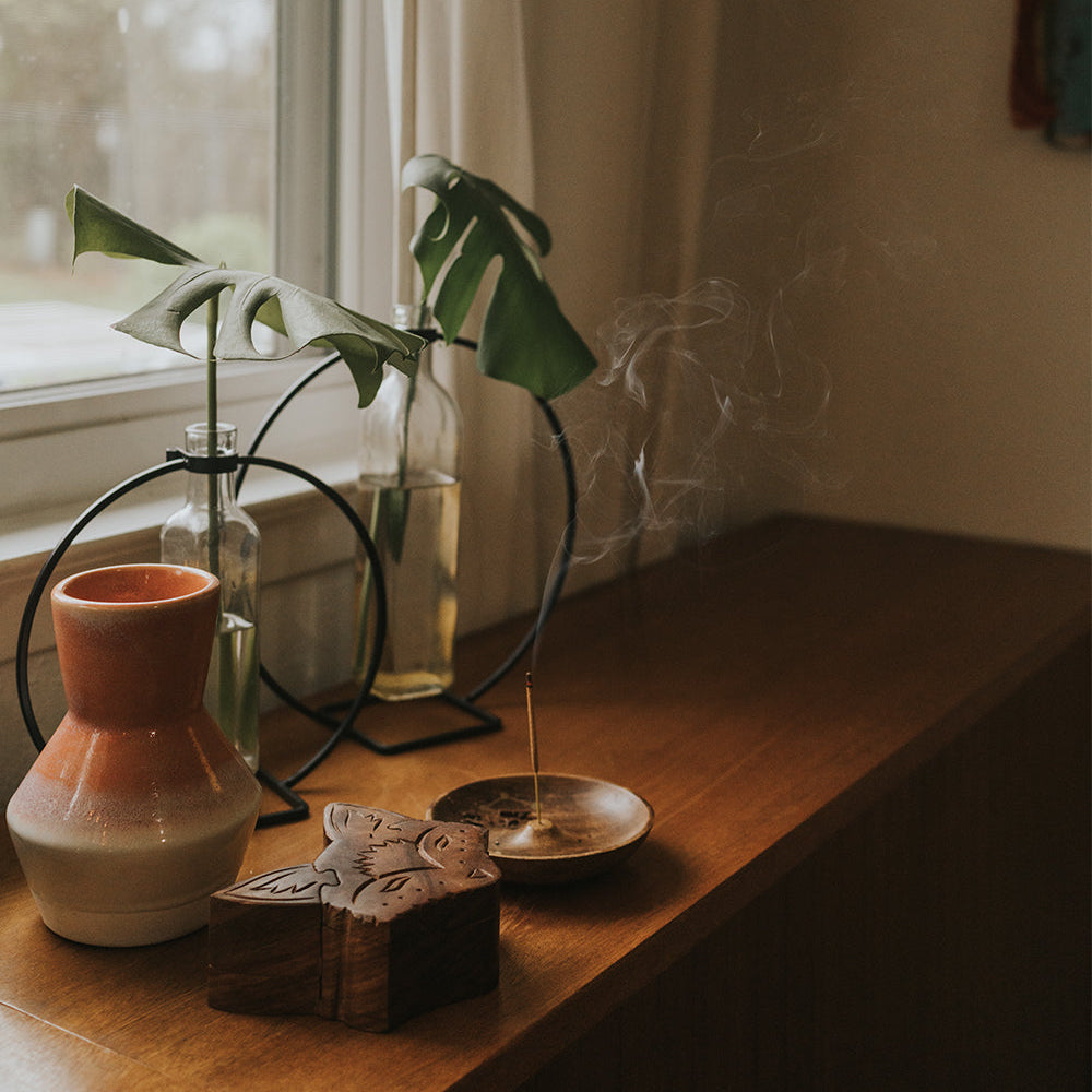 Jyotisha incense holder burning on a windowsill beside a vase and a carved wooden box