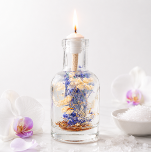 Cancer oil candle burning against a white background, blue and cream dried botanicals inside the glass, framed by a pale orchid bloom and a small bowl of coarse white sea salt.