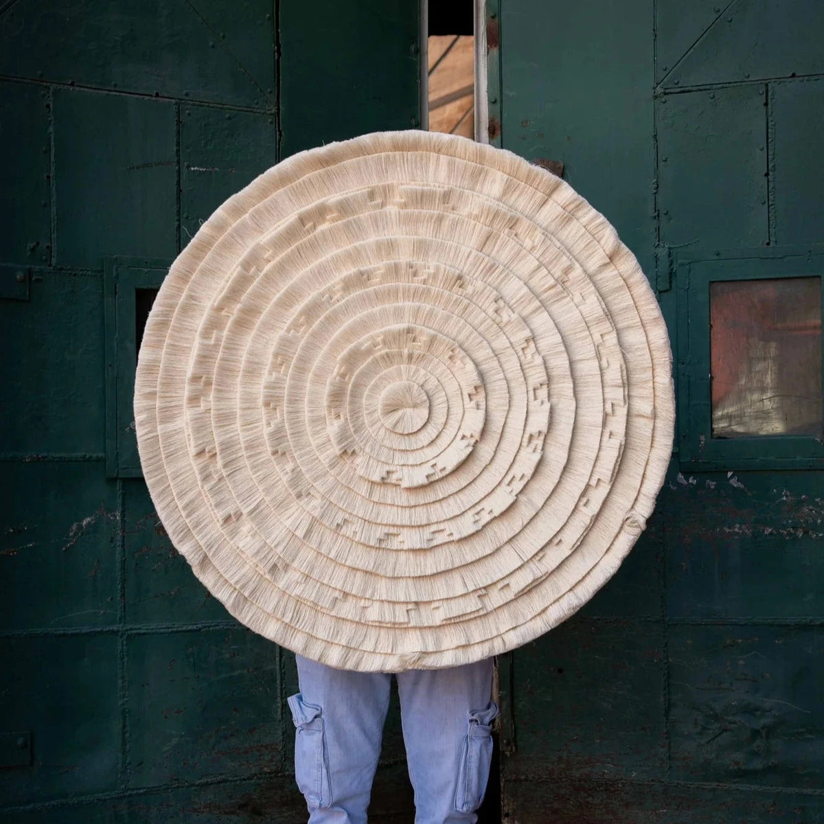 Person holding the round Calendario Azteca tapestry in front of a green metal door