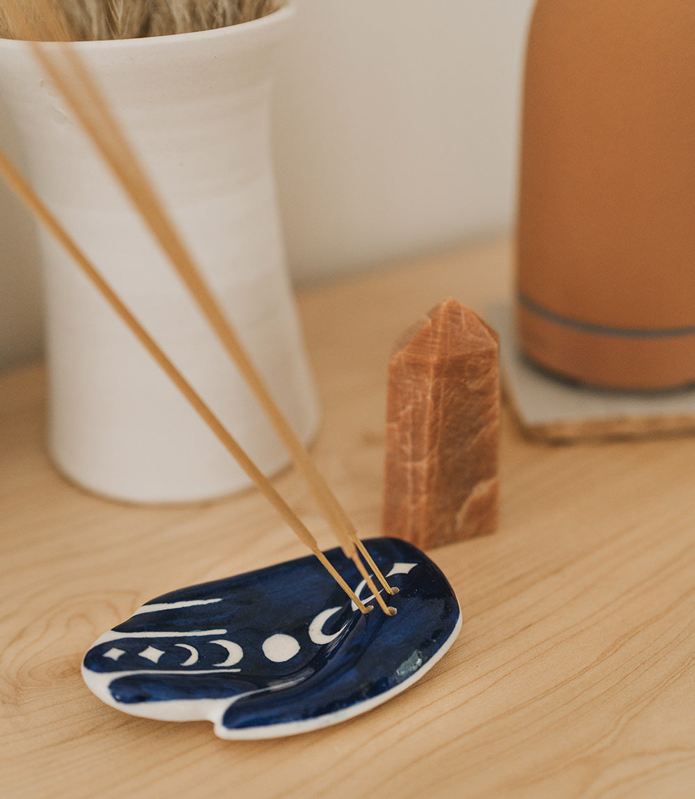 Jalini hamsa incense holder burning two sticks on a wooden table beside a crystal point