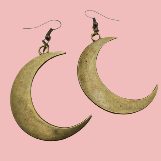 A pair of antiqued brass-tone crescent moon drop earrings on ear hooks, against a pink background.
