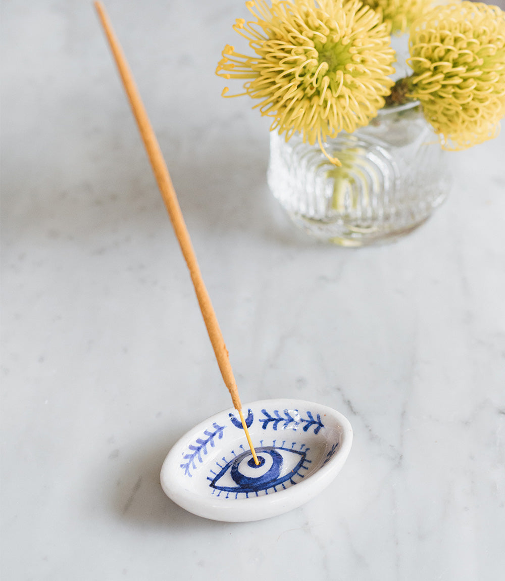 Evil eye incense dish burning a stick on marble beside a jar of yellow blooms