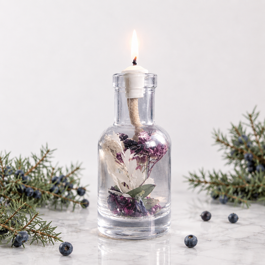 Lit Capricorn oil candle: a clear glass bottle of white dried flowers and juniper with a burning wick, on white marble beside juniper sprigs and dark blue juniper berries