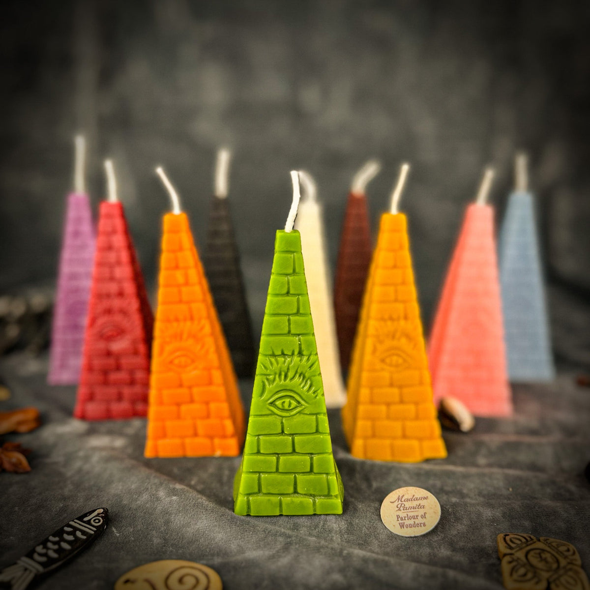 All ten colors of the pyramid candle grouped together on gray velvet, the green one in front and the Madame Pamita label beside it.
