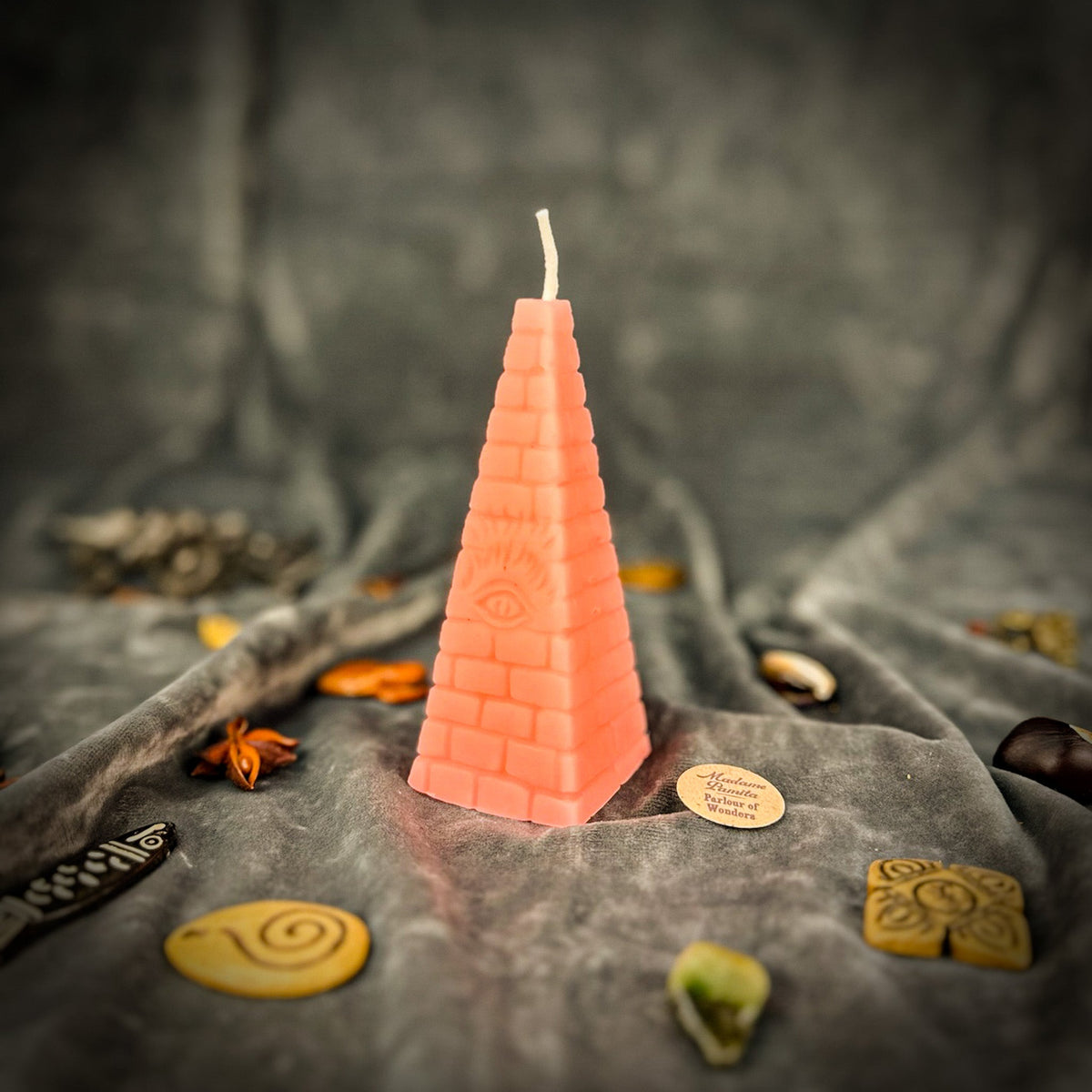 Coral-pink pyramid candle standing among charms and dried leaves on gray velvet.