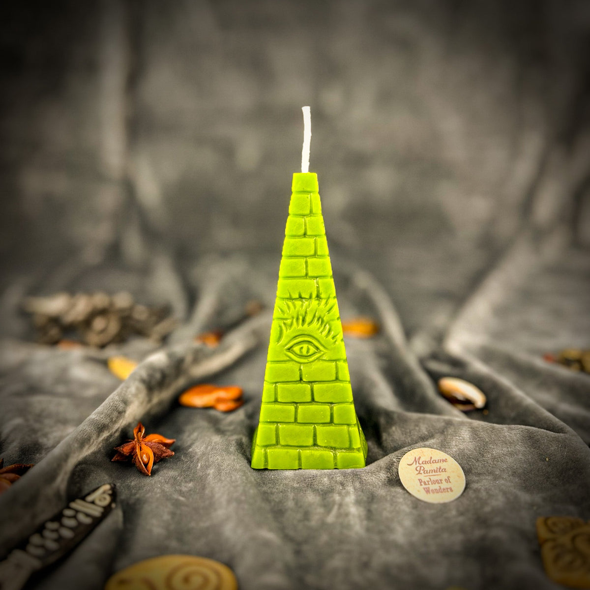 Green pyramid candle upright on gray velvet, the eye and its rays crisp above the lower courses of brick.