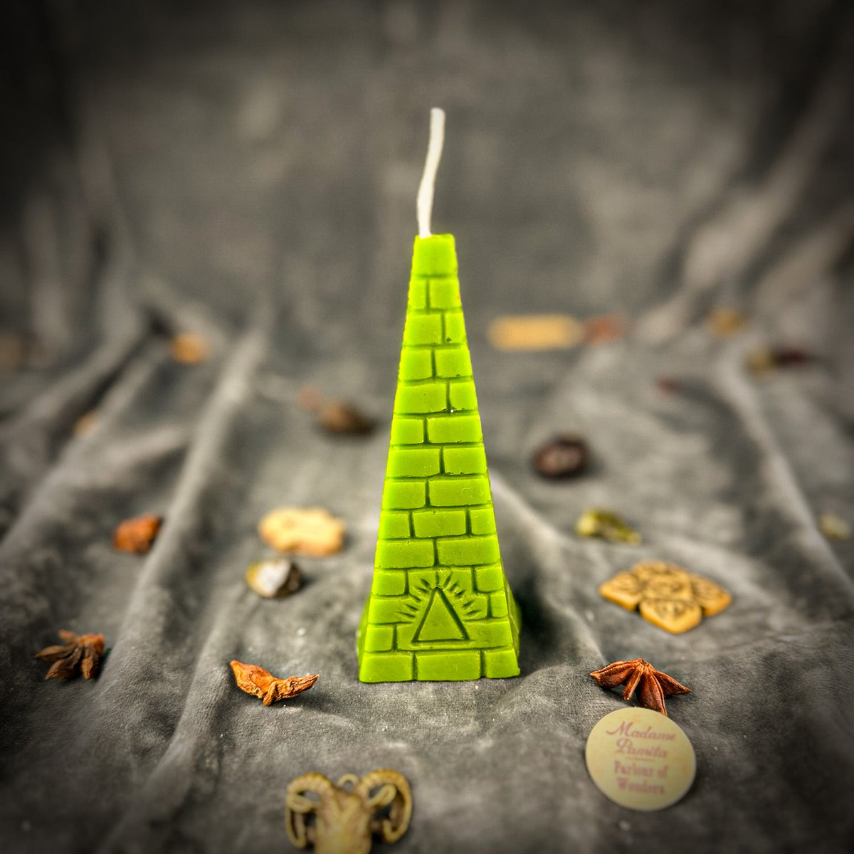 Green pyramid candle on gray velvet, this casting showing a rayed triangle rather than an open eye, surrounded by scattered charms and star anise.