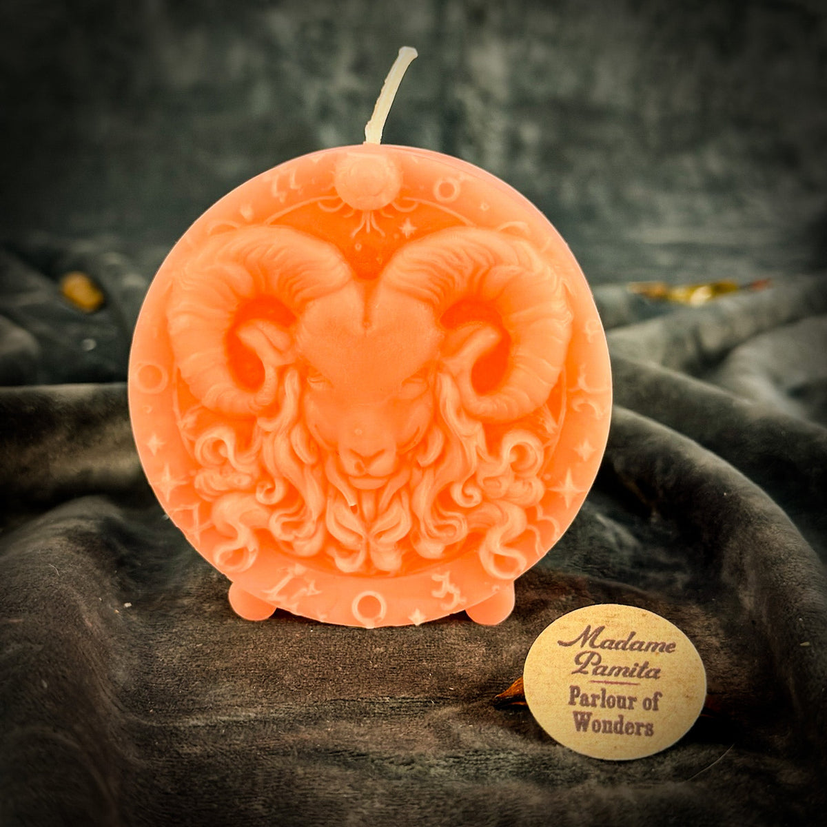 Coral-orange Aries medallion candle, wick unlit above the ram's head.