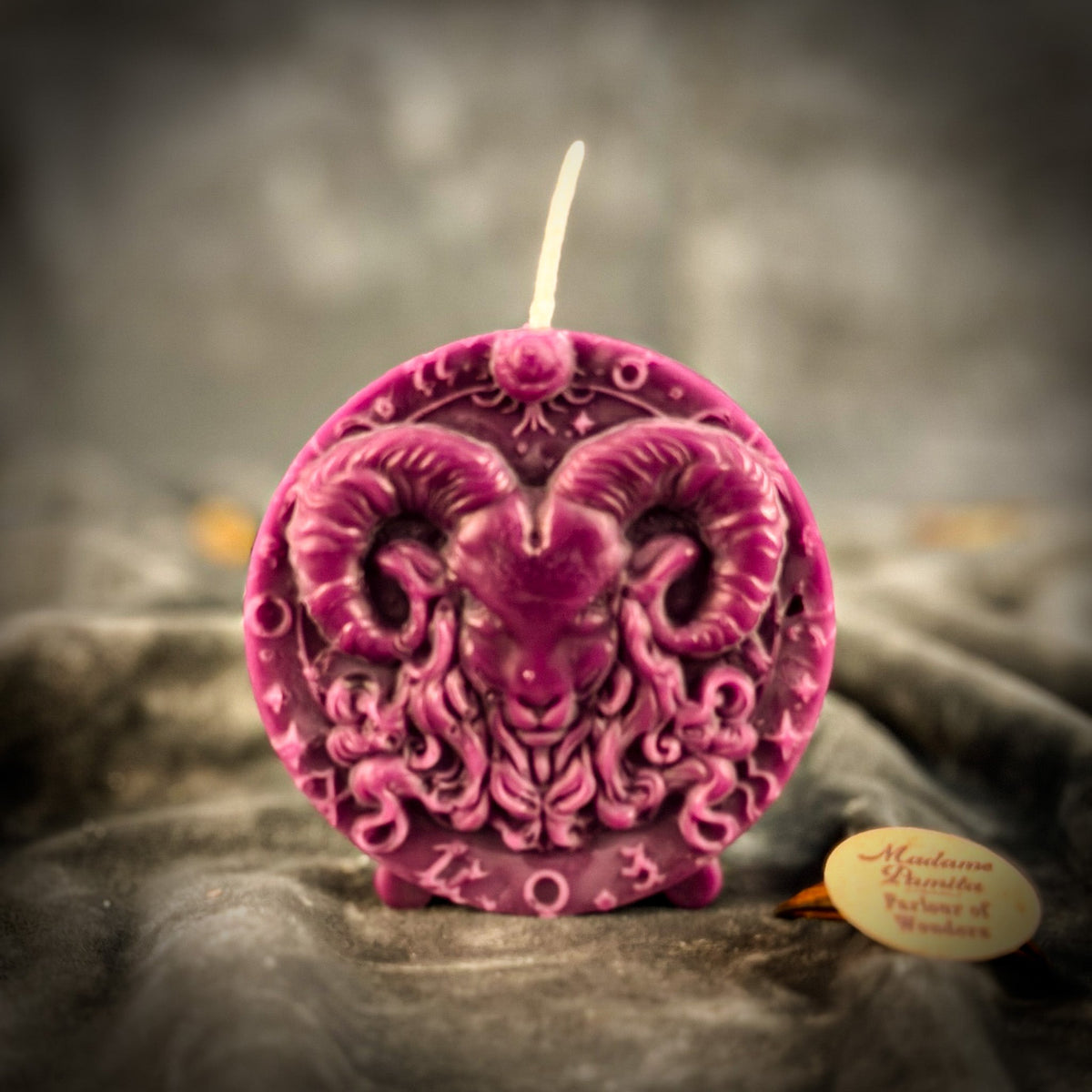 Magenta Aries medallion candle standing on gray velvet, glyphs legible along the lower rim.