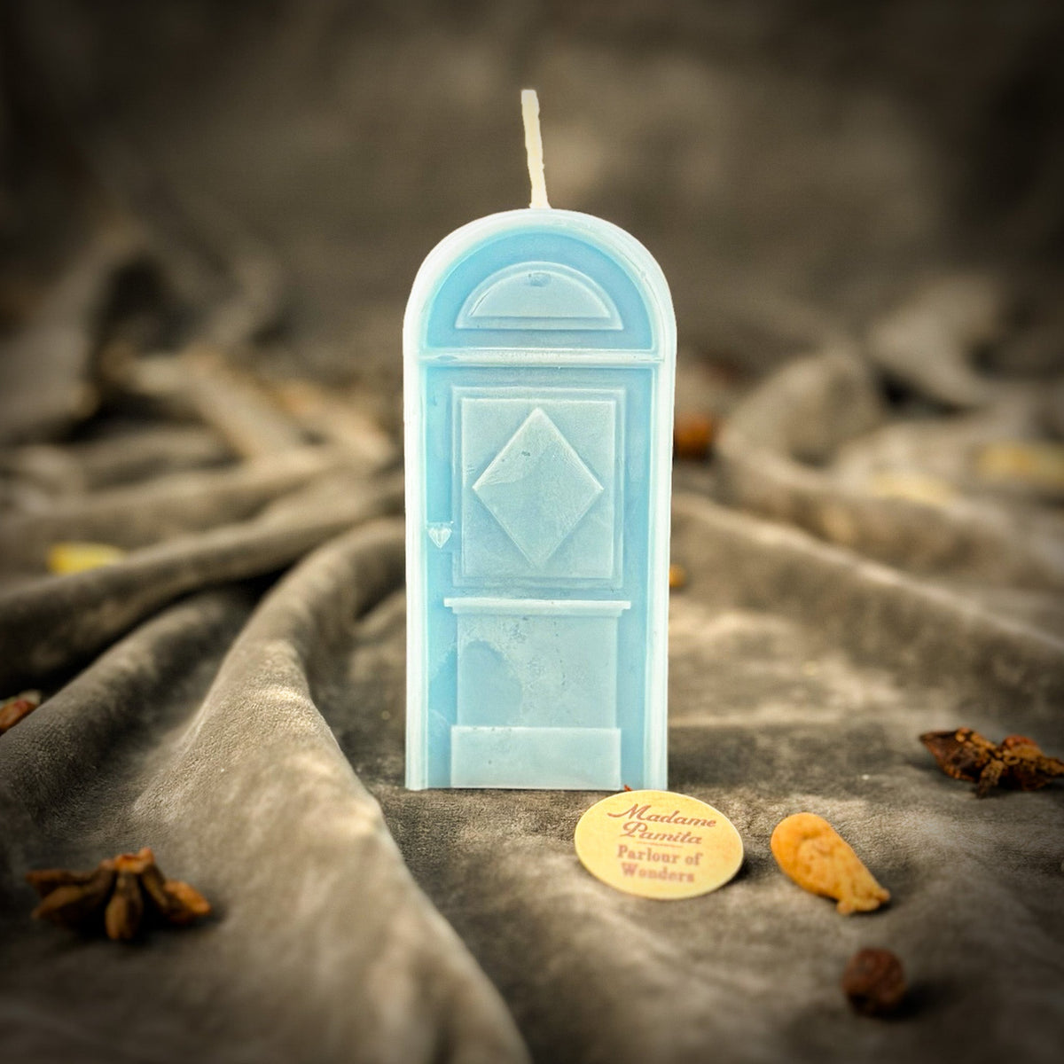 Pale blue door-shaped spell candle standing on draped gray velvet with the maker's token and whole spices beside it