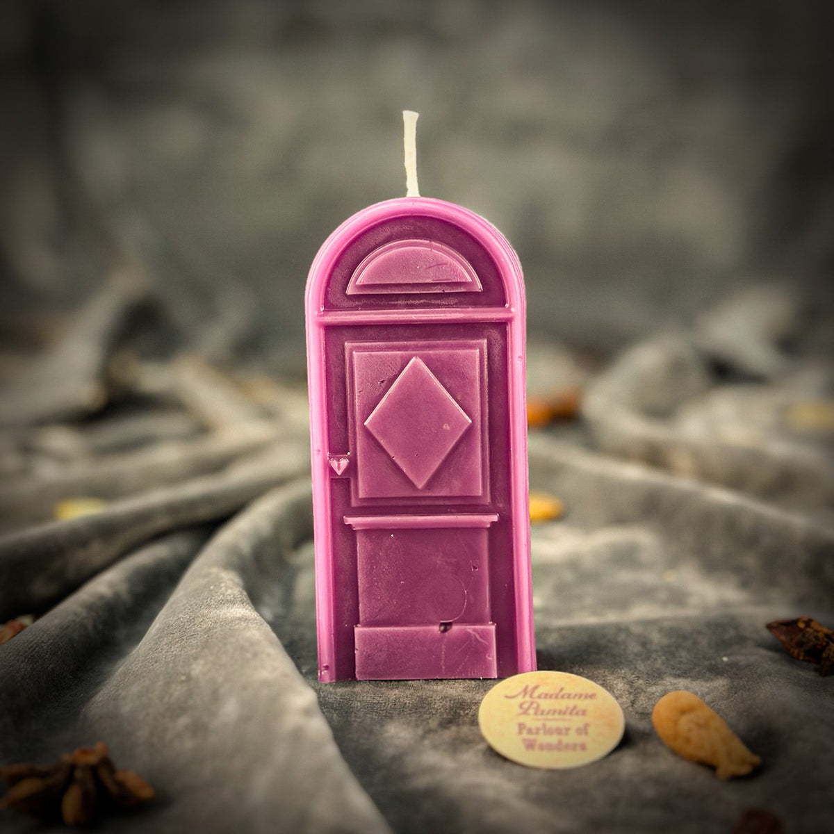 Purple door-shaped spell candle on gray velvet with the round Madame Pamita Parlour of Wonders token and scattered star anise