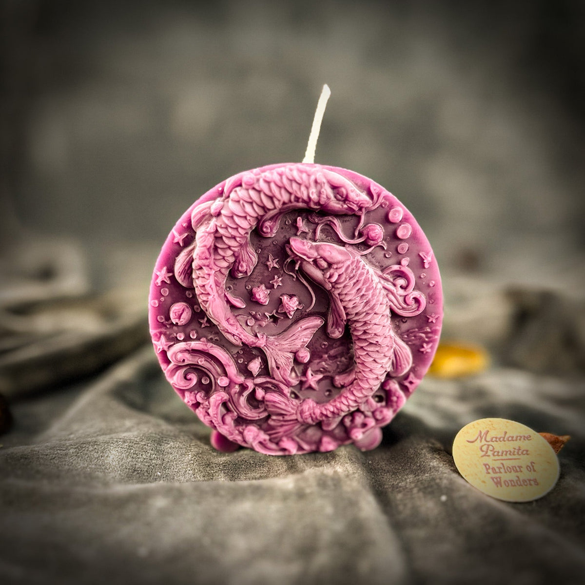 Rose-pink Pisces medallion candle standing on gray velvet.