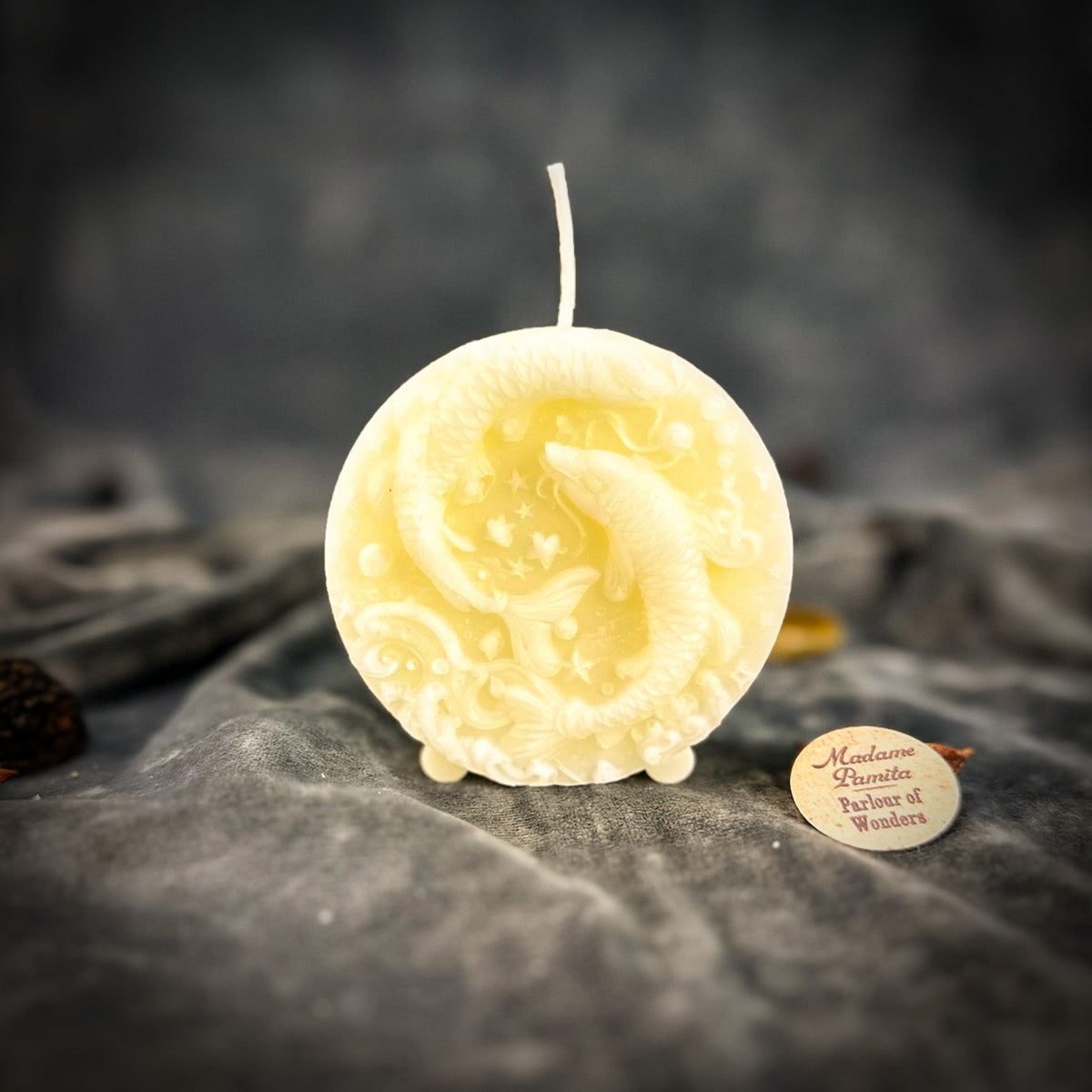 Cream-white Pisces medallion candle on gray velvet, the wave scrollwork soft in the pale wax.