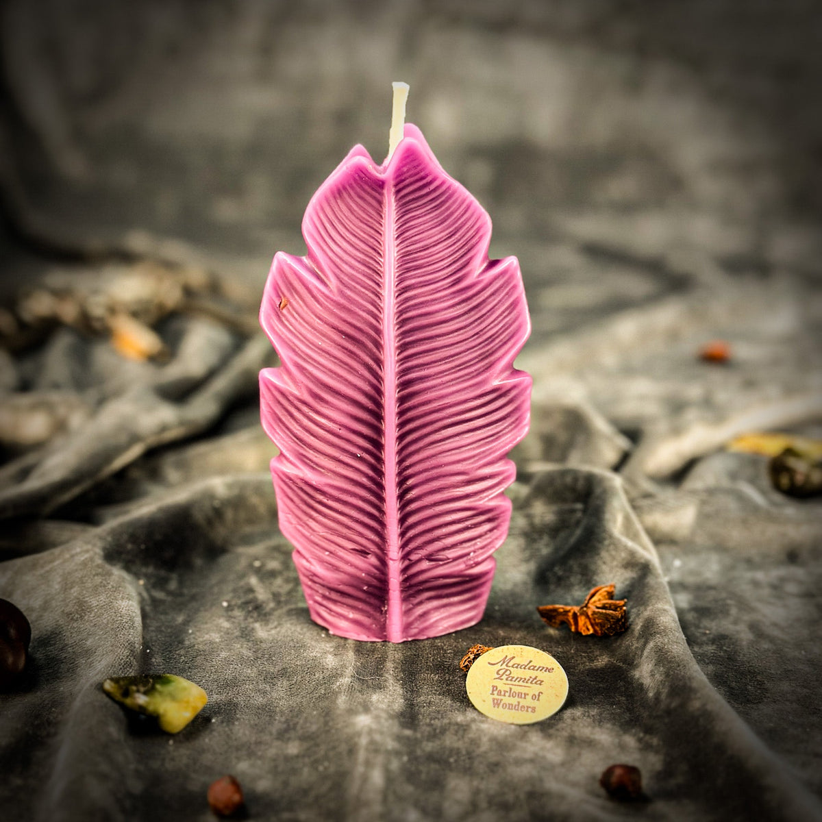 Magenta leaf-shaped candle standing on gray velvet, the maker's token beside it