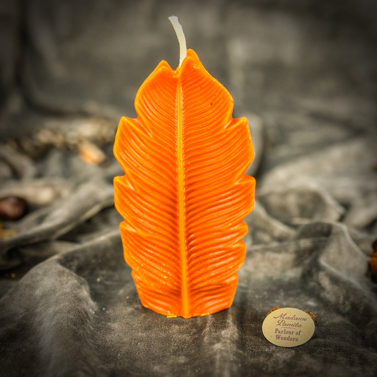 Bright orange leaf-shaped candle standing on gray velvet with the Parlour of Wonders token in front