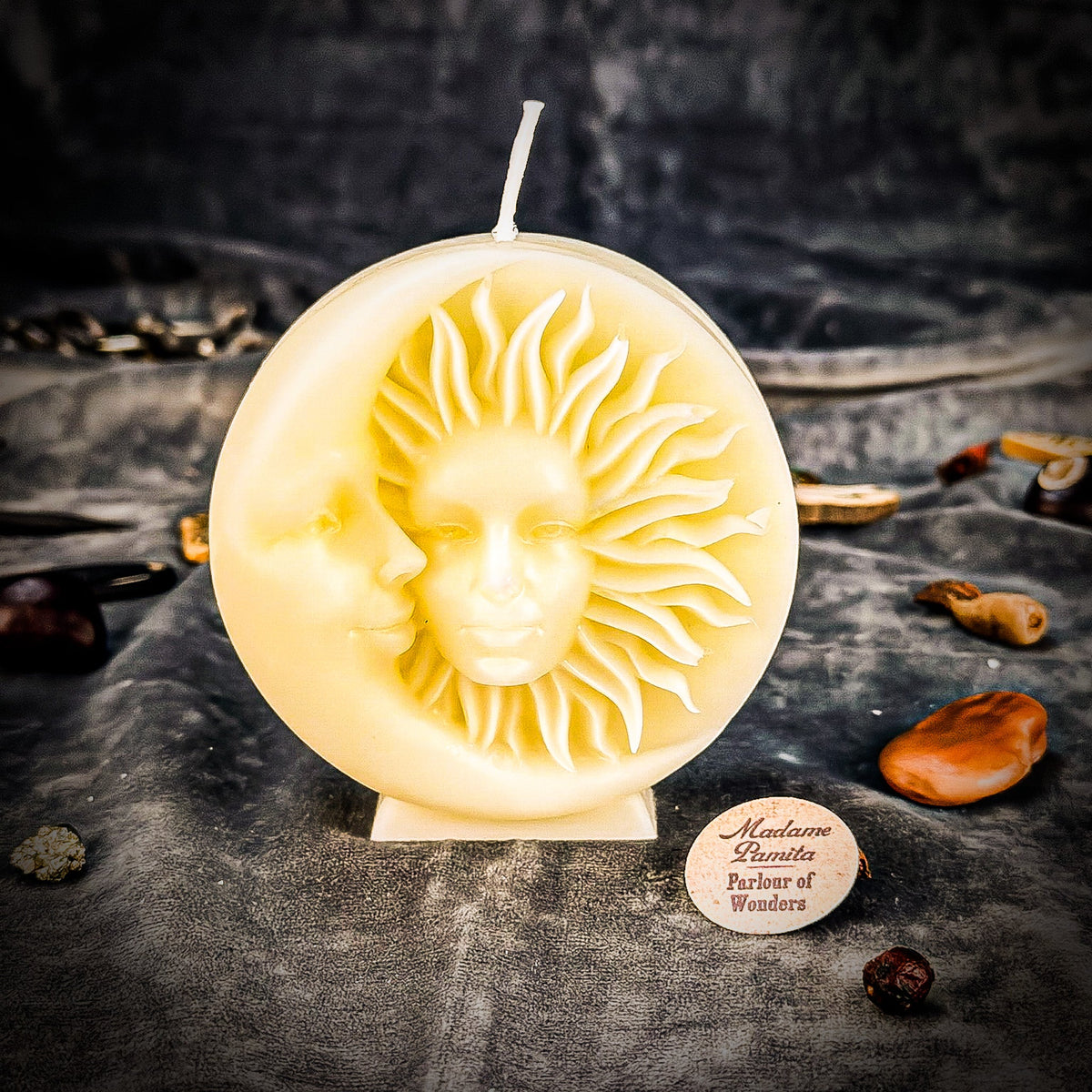 Cream sun-and-moon disc candle on gray velvet, the moon profile and radiating sun face standing out in pale relief