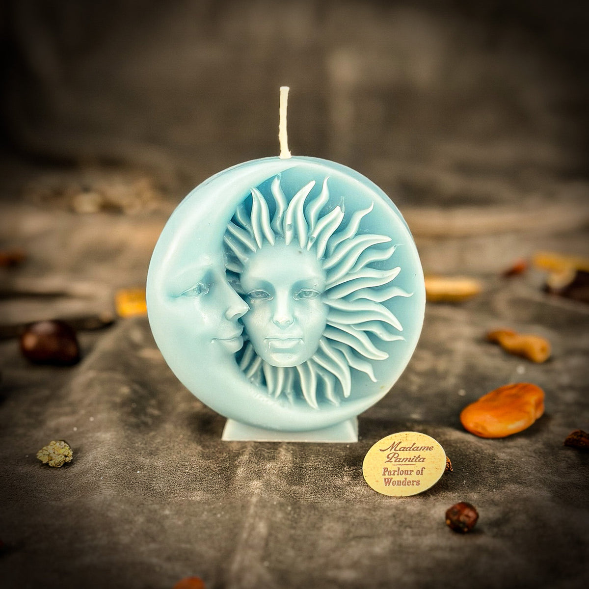Pale blue sun-and-moon disc candle on gray velvet, the sun's rays picked out in soft relief