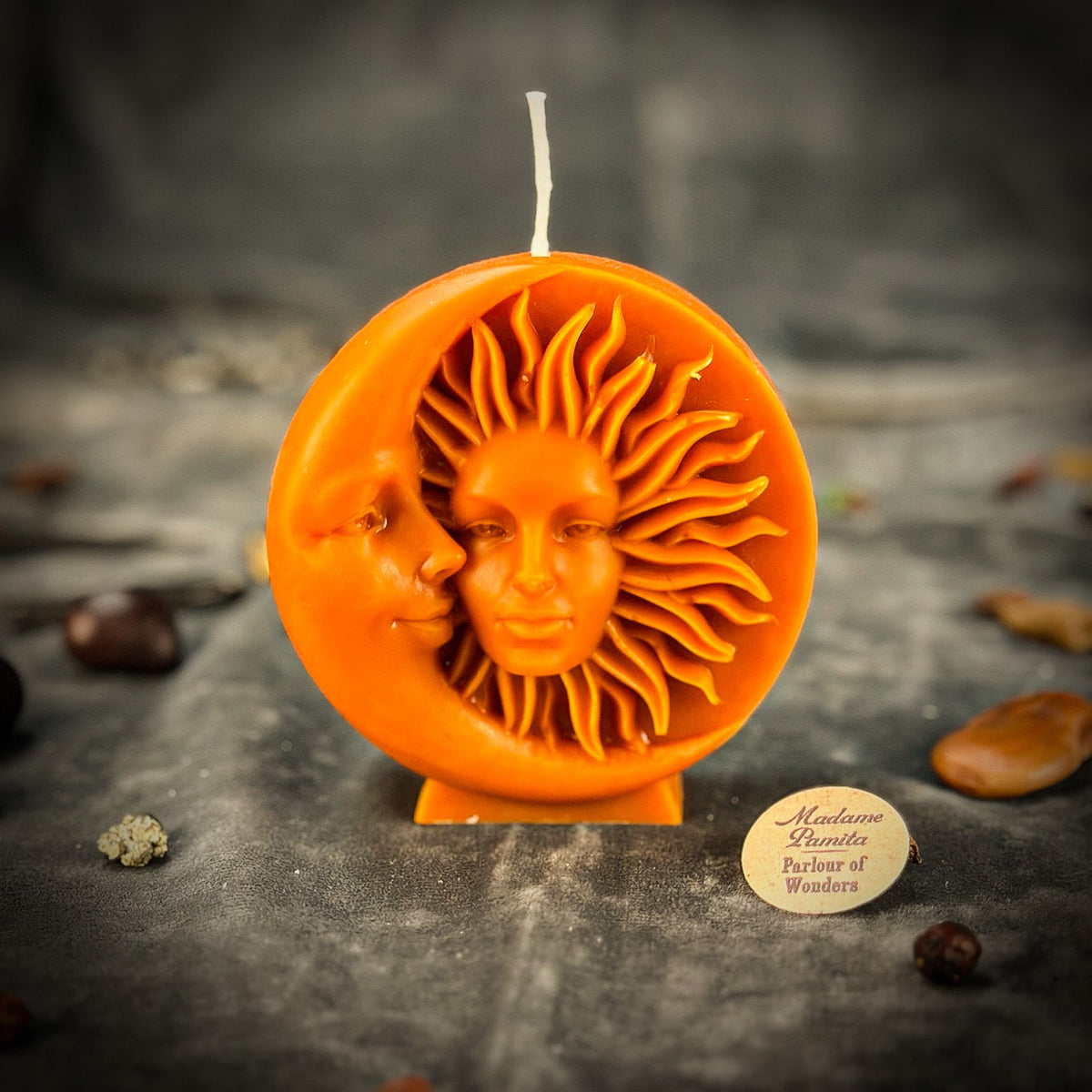 Orange sun-and-moon disc candle on gray velvet with whole spices and seeds around the base