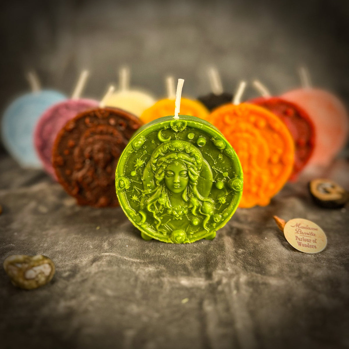 All ten colors of the Virgo medallion candle grouped on gray velvet, the green one in front beside the Madame Pamita label.