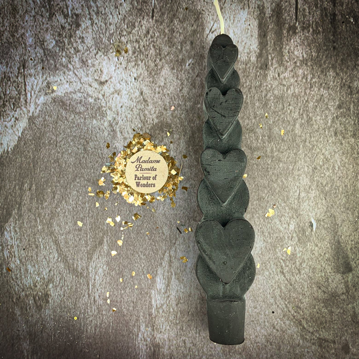 The Black (Protection) Love Spell Heart Taper in dark charcoal beeswax on a mottled stone backdrop, the gold-flecked Madame Pamita token beside it.