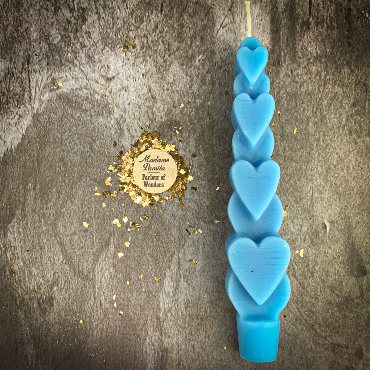 The Blue (Reconciliation) Love Spell Heart Taper in bright blue beeswax on a mottled stone backdrop, the gold-flecked Madame Pamita token beside it.
