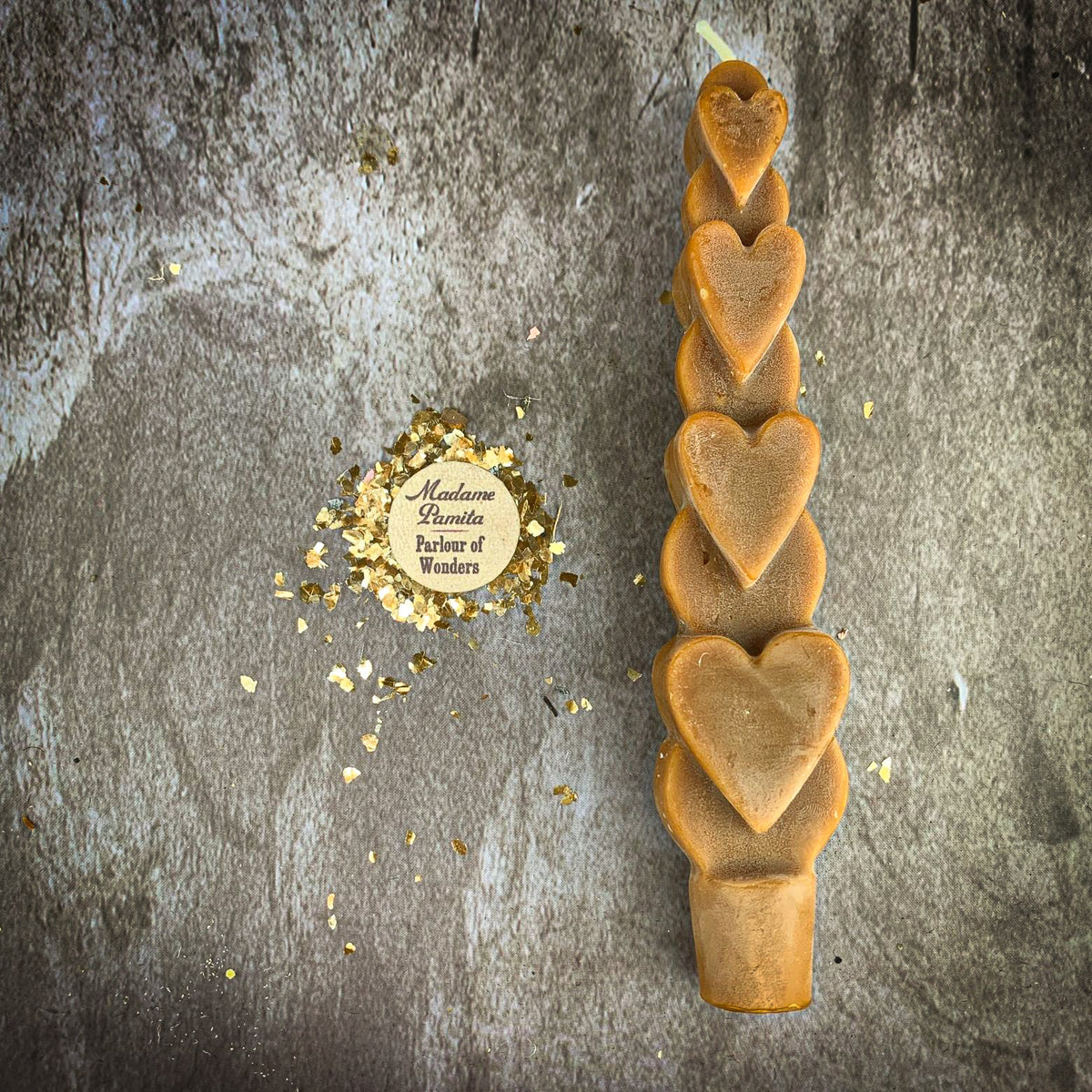 The Brown (Grounding and Fidelity) Love Spell Heart Taper in caramel beeswax on a mottled stone backdrop, the gold-flecked Madame Pamita token beside it.