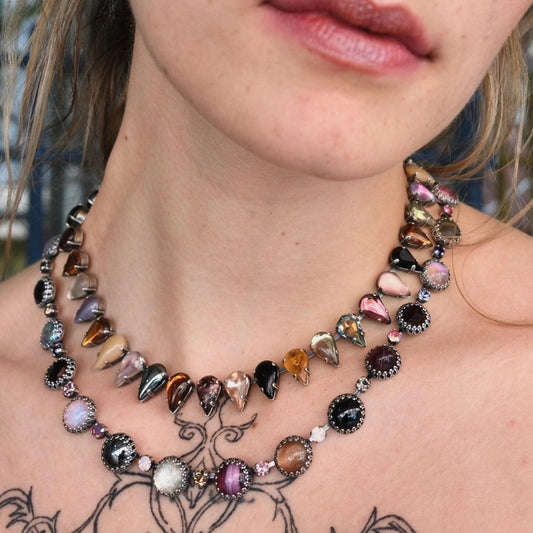 The fang choker worn at the collarbone over a chest tattoo, its teardrop glass stones alternating with round bezel-set stones.