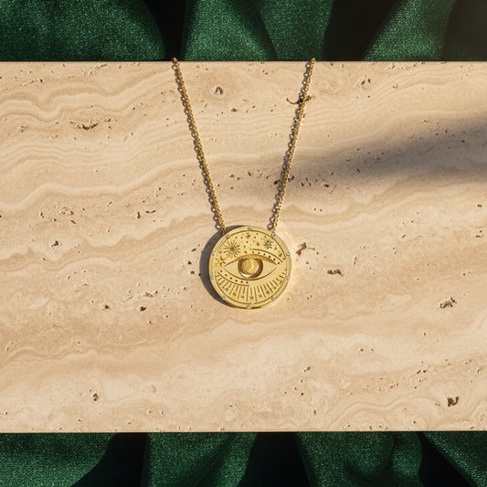 A gold-tone disc pendant with an eye motif resting on travertine stone beside green velvet.