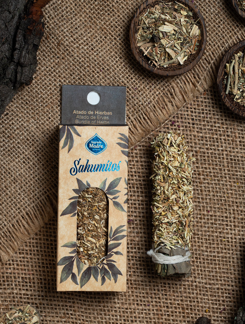Boxed smudge stick beside a white sage bundle and bowls of loose herbs on burlap