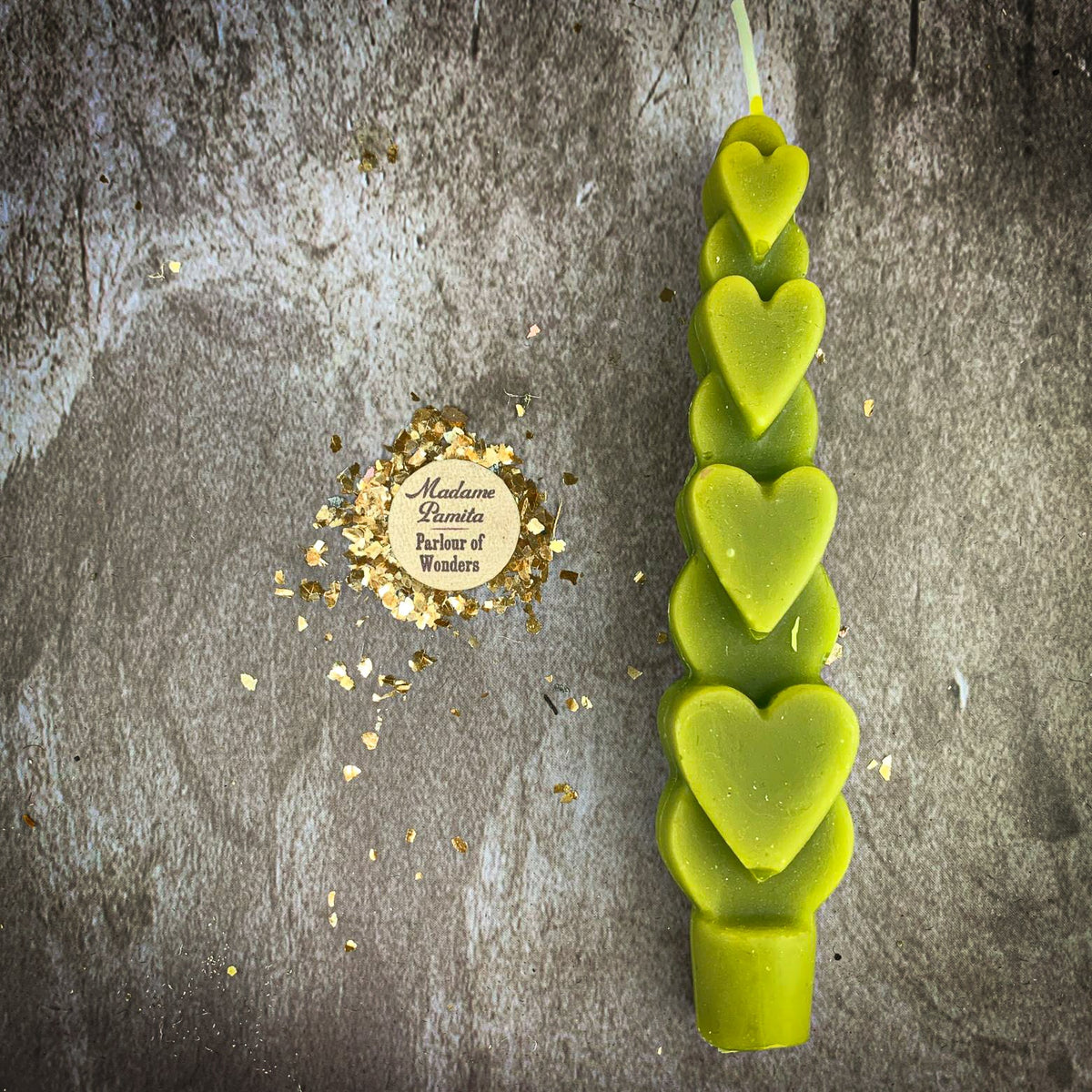 The Green (Generosity) Love Spell Heart Taper in olive-green beeswax on a mottled stone backdrop, the gold-flecked Madame Pamita token beside it.