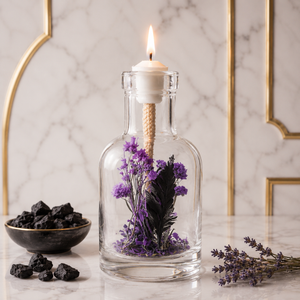 Lit Lilith oil candle: a clear glass bottle of deep purple and near-black dried flowers with a burning wick, on white marble against a brass arch, beside a dark bowl of black resin and cut lavender