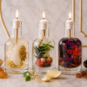Three lit Oracle Trio oil candles side by side on marble before a brass arch: clear glass bottles holding golden wheat, green fern with strawberry, and deep red botanicals, each with a burning wick