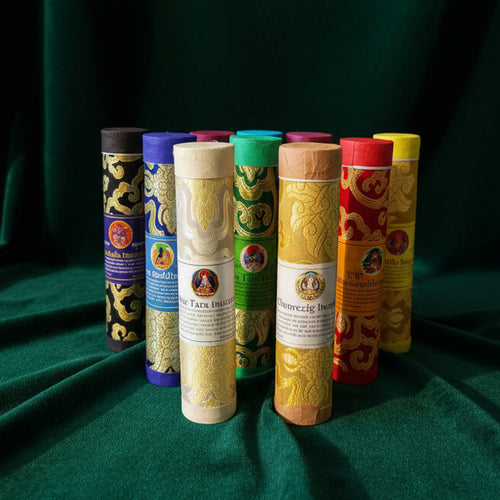 Ten brocade-wrapped incense tubes standing together on green velvet — black, blue, magenta, green, cream, orange, red and yellow, each with its own printed label.