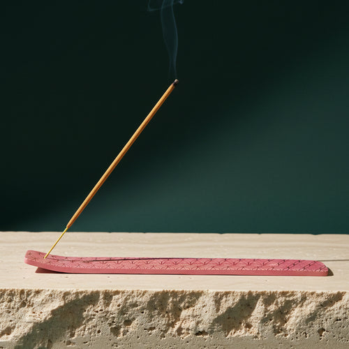 Incense burner in a pink finish on a stone ledge against a dark green backdrop, a lit stick sending up a thin line of smoke
