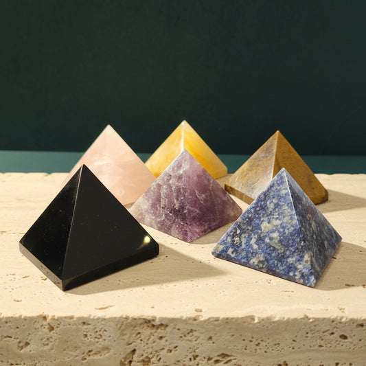 Six polished crystal pyramids on a travertine slab against a dark green backdrop: black obsidian, rose quartz, warm yellow, purple, tiger eye and mottled blue