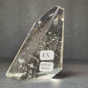 Tall smoky quartz free-form with a slanted polished tip, dense mineral inclusions clouding the lower half, carrying the EX and Made in Brazil labels