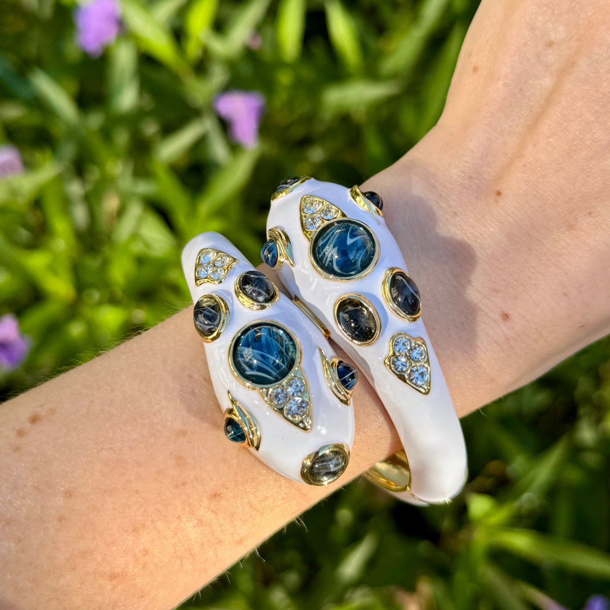 The white enamel snake bracelet worn on a wrist against green foliage, the two heads sitting side by side across the top of the wrist.