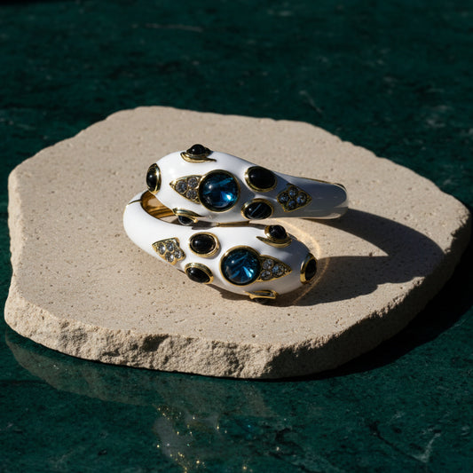 White enamel double-headed snake bracelet on a pale stone slab over dark green, the two heads facing each other, each set with a domed blue cabochon and clear pave accents in gold-tone bezels.