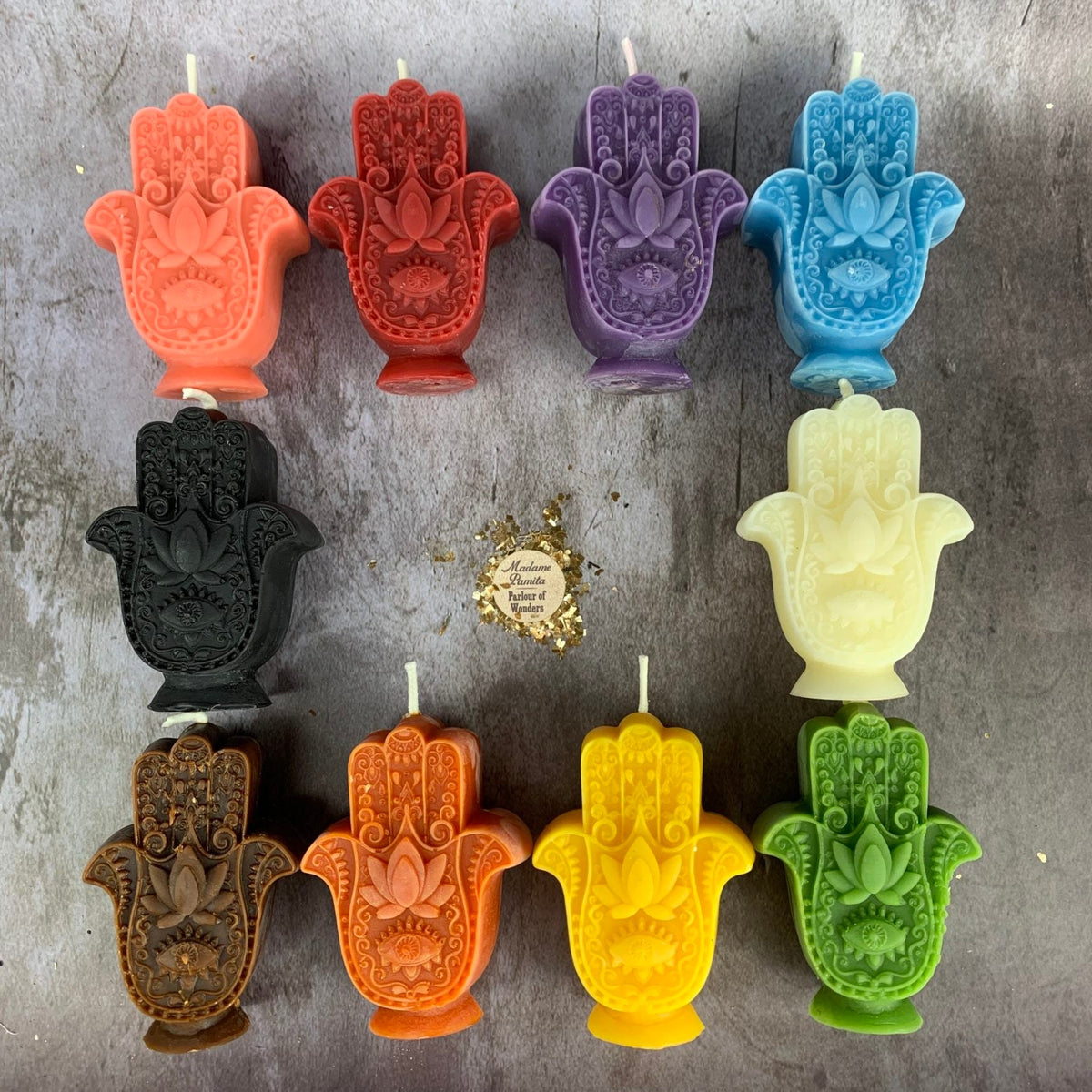 All ten colors of the Hamsa candle laid out on gray concrete — coral, red, purple and blue on top; black and cream in the middle; brown, orange, yellow and green below.