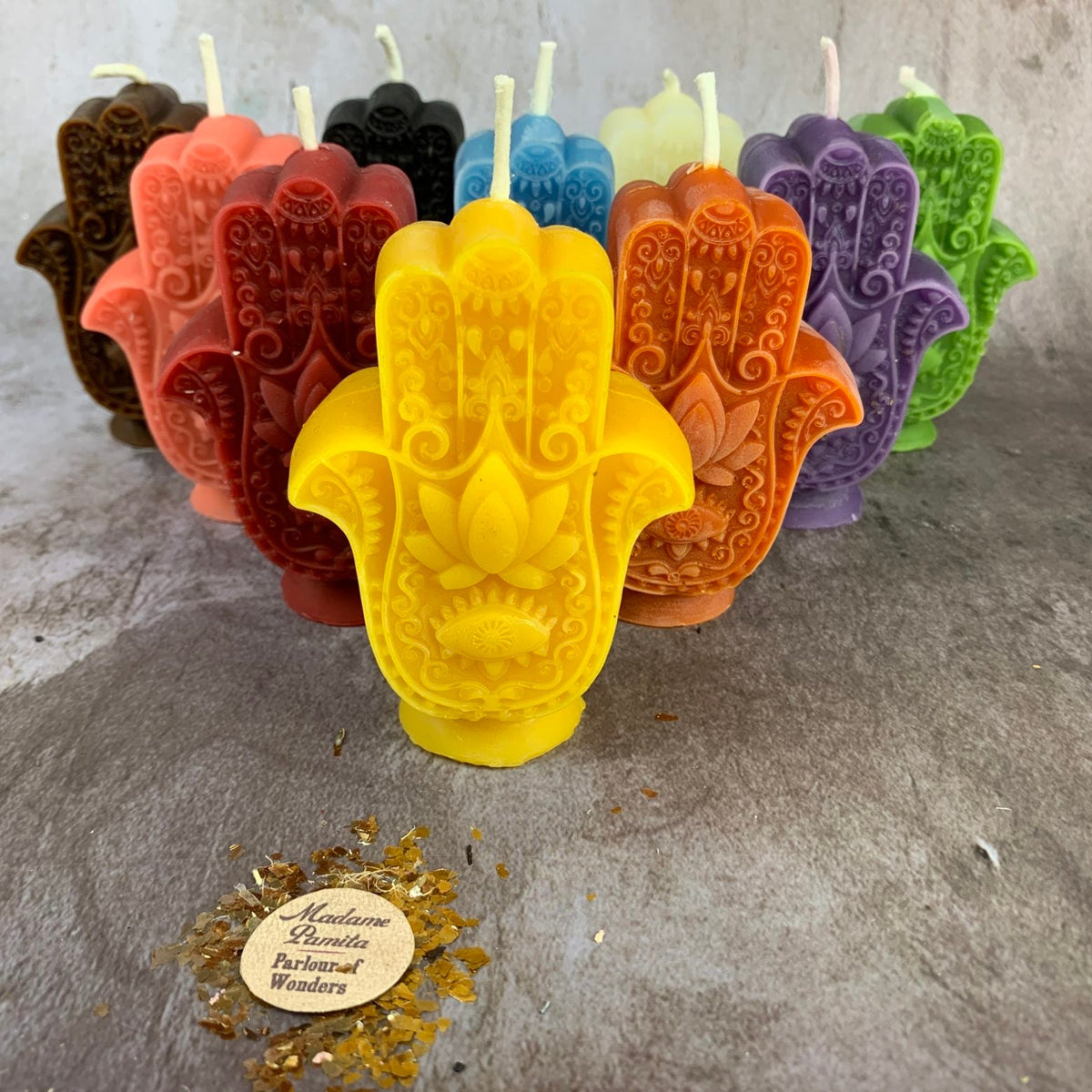 Group of Hamsa candles in every color standing behind a yellow one, with the Madame Pamita wax-seal label and gold leaf flakes in front.