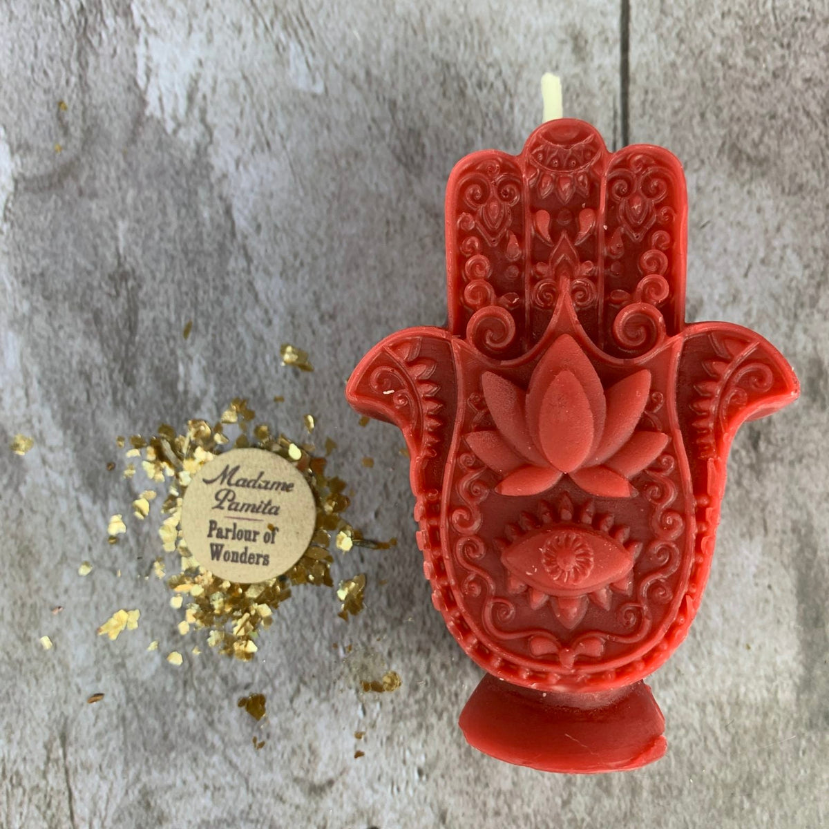 Red Hamsa candle beside the round Madame Pamita label and scattered gold leaf.