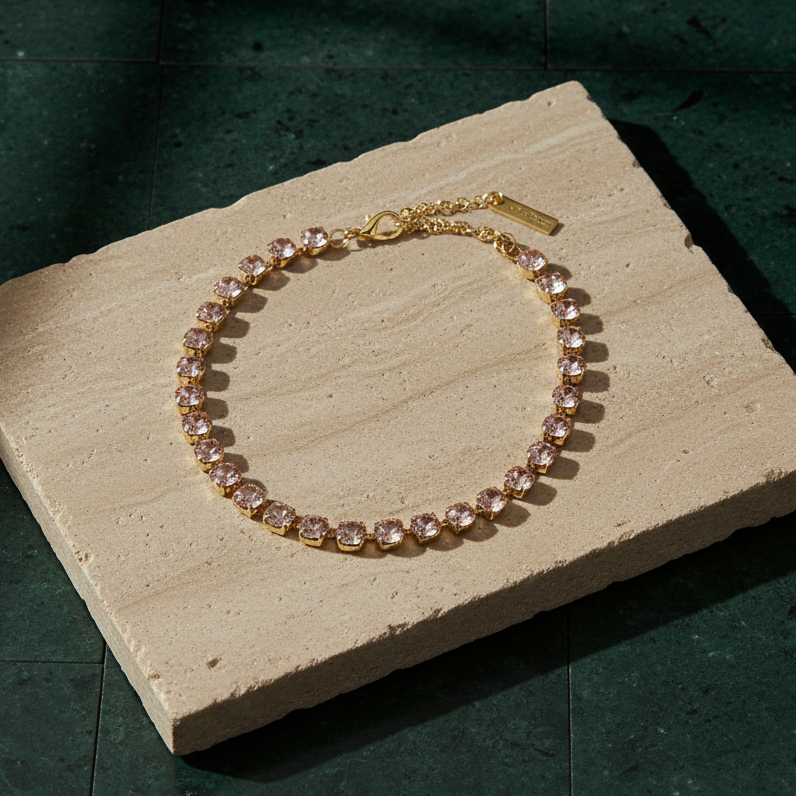 Single-strand necklace of round pale pink crystals in gold-tone settings, laid on a stone slab with its extension chain and branded tag.