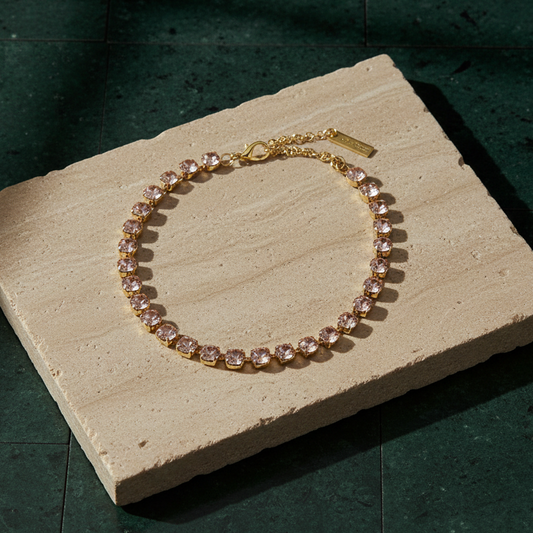 Single-strand necklace of round pale pink crystals in gold-tone settings, laid on a stone slab with its extension chain and branded tag.