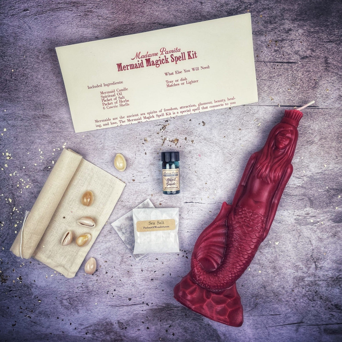 Deep red mermaid candle with the kit, the instruction card listing candle, oil, salt, herbs and six cowrie shells.