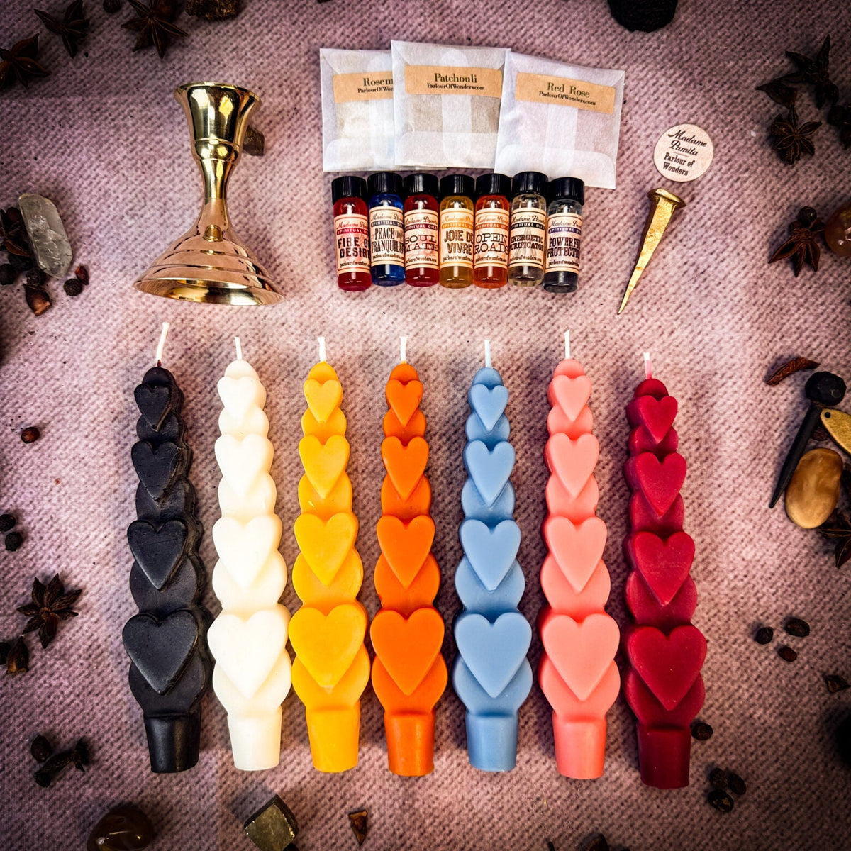 The full range on pink cloth: seven heart-stacked taper candles in black, white, yellow, orange, blue, pink and red, with a brass holder, seven oil vials, three herb sachets and a brass snuffer