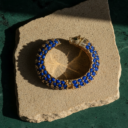 Navy blue panther cuff on a pale stone slab, rows of blue oval cabochons with gold beading and a pavé-set panther head.