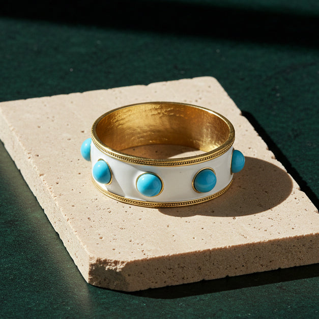 Wide white enamel bangle on a pale stone slab, gold beaded edges and round turquoise-blue glass cabochons, with a gold interior.