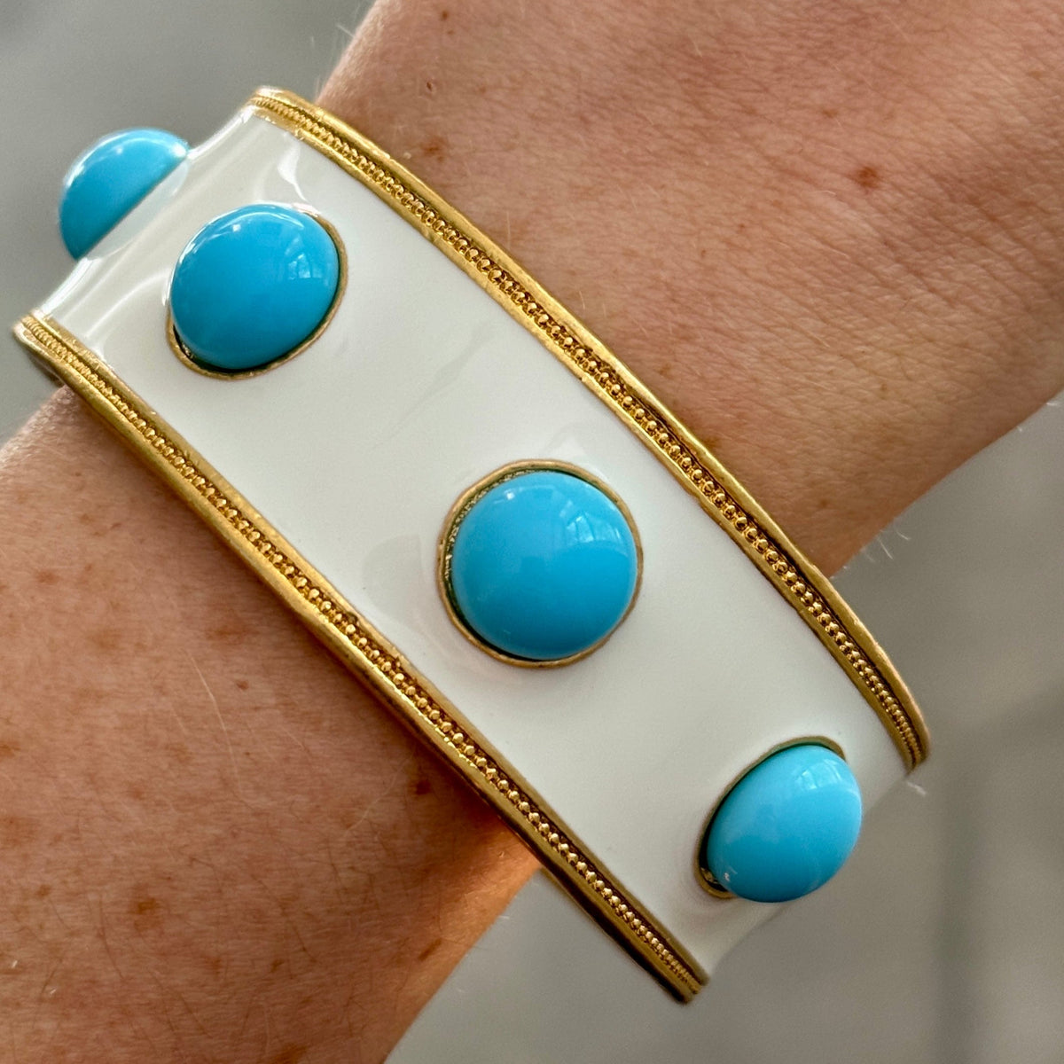 The white enamel bangle worn on a wrist, four round turquoise-blue glass cabochons visible along the band.