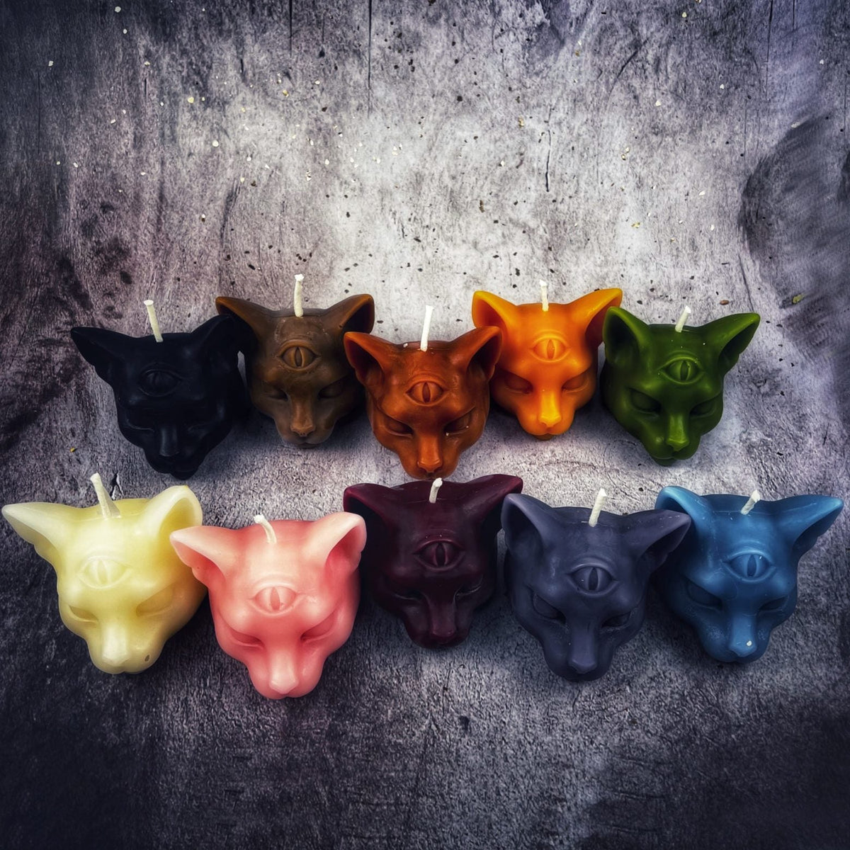 All ten colors of the cat head candle in two rows on gray stone — black, brown, rust, orange and green above; pale yellow, salmon, wine, navy and teal below.